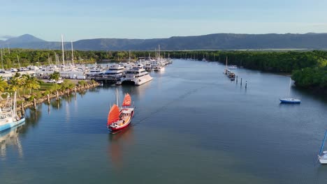 aerial footage captures a red-sailed tour boat cruising into a marina surrounded by yachts and lush greenery, under soft golden hour lighting