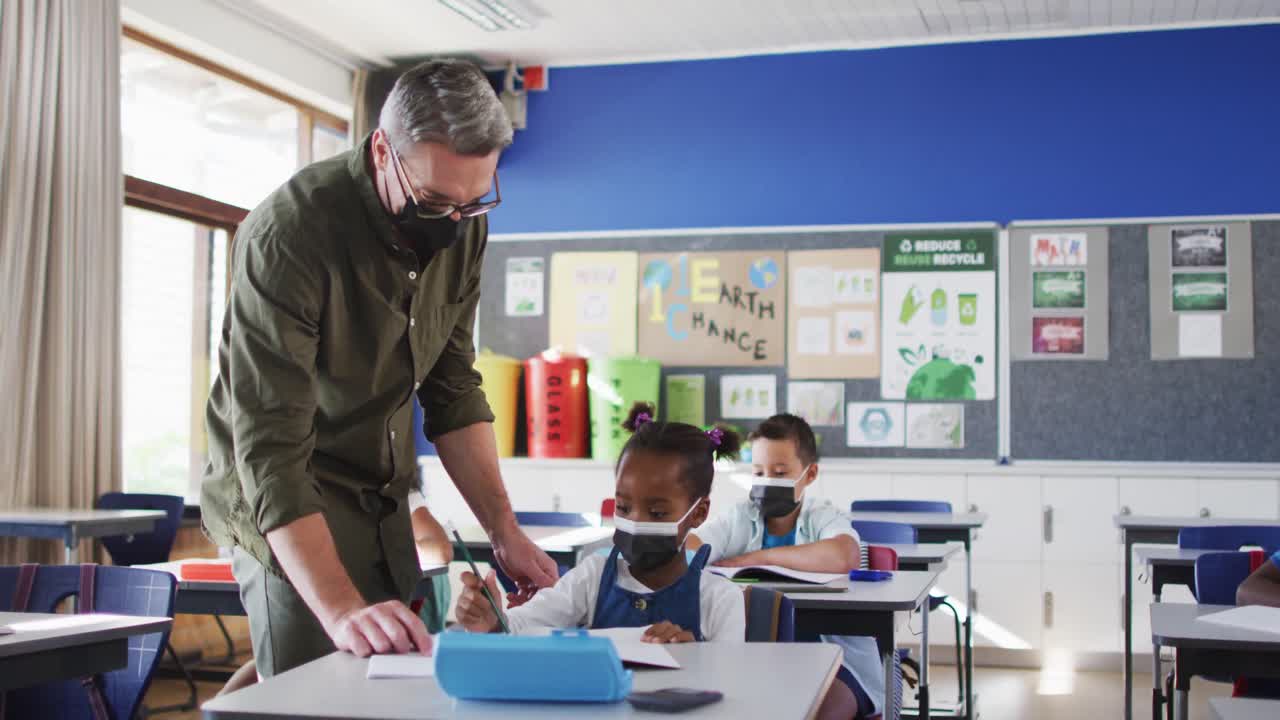 un maestro masculino diverso ayudando a una estudiante sentada en el aula, todos con máscaras faciales