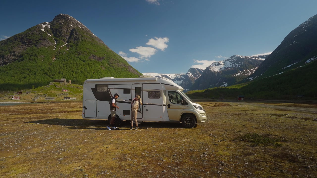 Escena estática de una pareja saliendo de su autocaravana, poniéndose los zapatos y caminando hacia un impresionante paisaje montañoso durante una aventura de viaje.