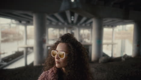 curly-haired woman in yellow sunglasses looking to side under urban overpass