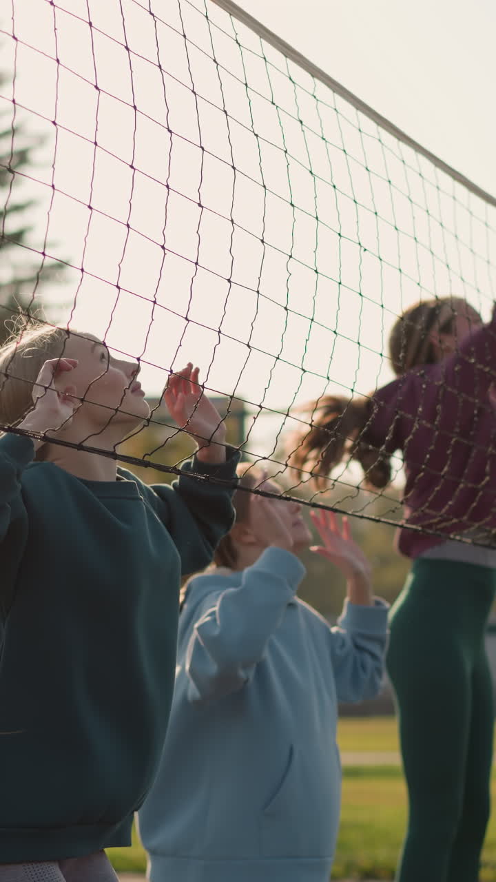 후디를 입은 여성들이 코트 밖에서 배구 훈련에 참여합니다. 배경 특징, 초록색, 그리고 건물, 여성들은 연습 세션 중에 손을 들고 뛰어내립니다.