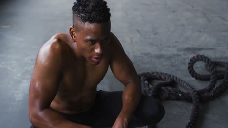 hombre afroamericano sin camisa descansando después de luchar con cuerdas en un edificio urbano vacío