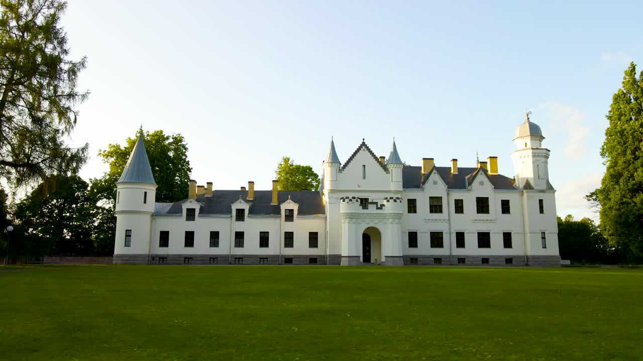 Low angle wide shot footage gliding close to green grassy ground in front of a white large and majestic Alatskivi castle during a summer cloudy and sunny evening or day. Green trees and gorgeous yard.