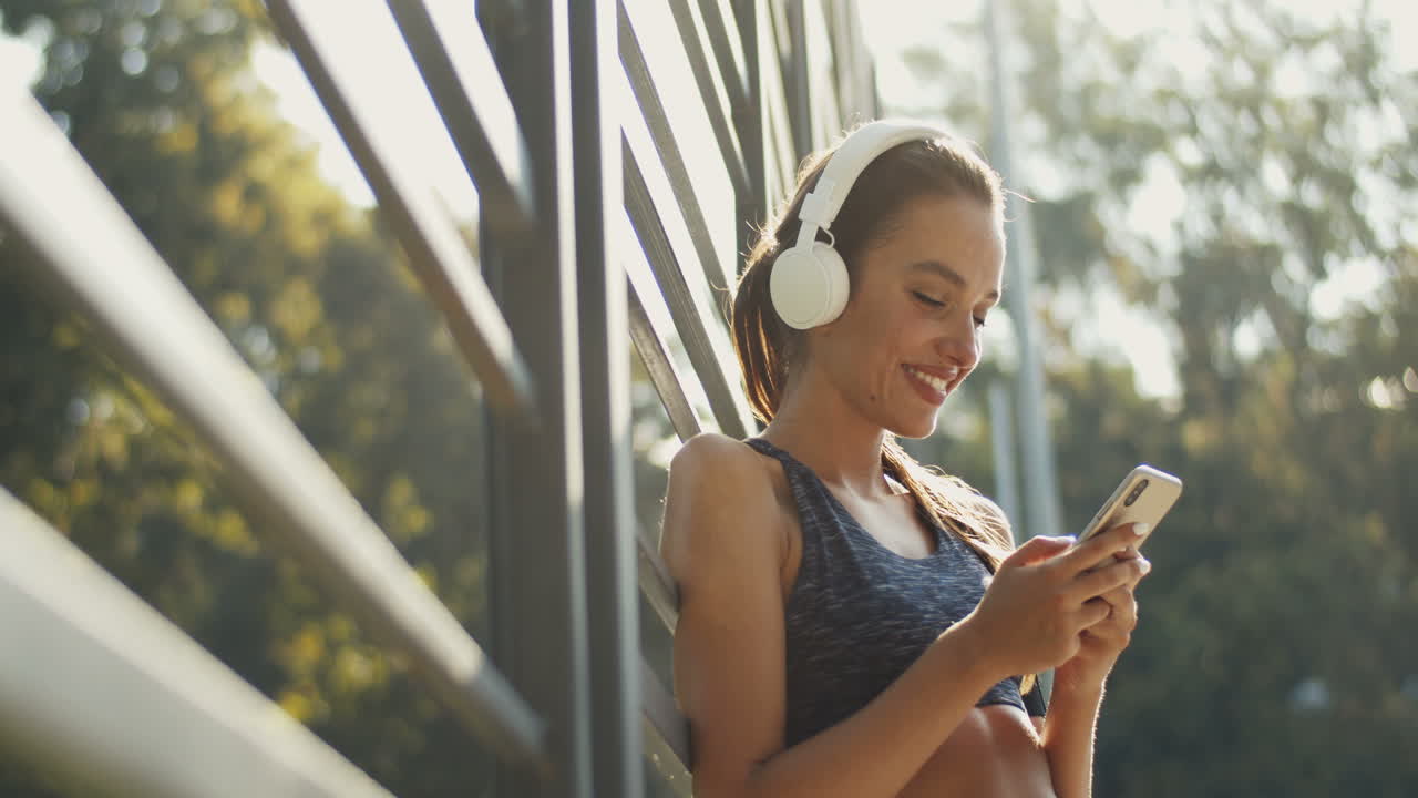 chica deportiva con mensajes de texto de auriculares en el teléfono inteligente y riéndose de la cancha al aire libre en un día de verano