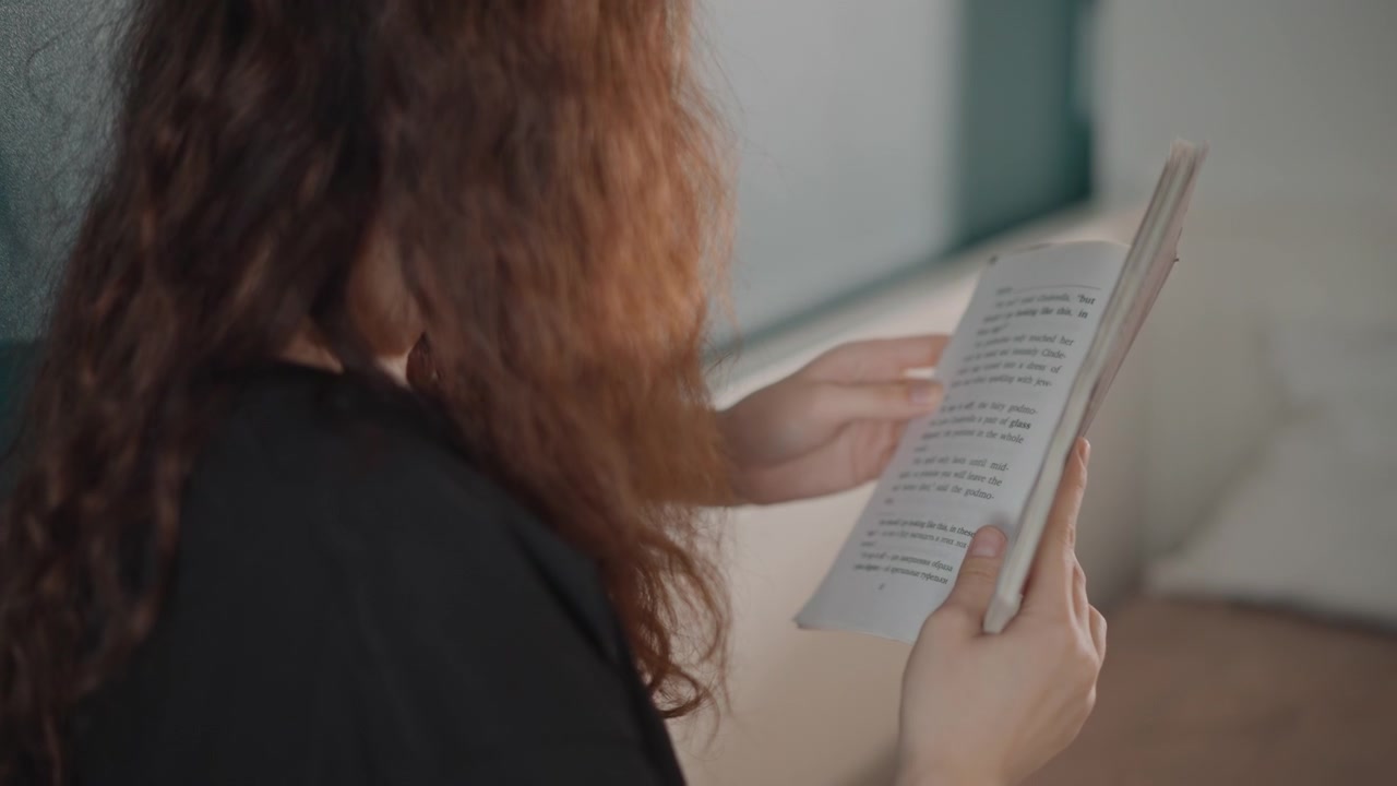 Vista próxima por cima do ombro de uma jovem mulher com cabelo longo e encaracolado, folheando um livreto, com as mãos no papel enquanto lê com expressão concentrada. Uma parede azul-petróleo e uma luz lateral suave criam um ambiente interno tranquilo.