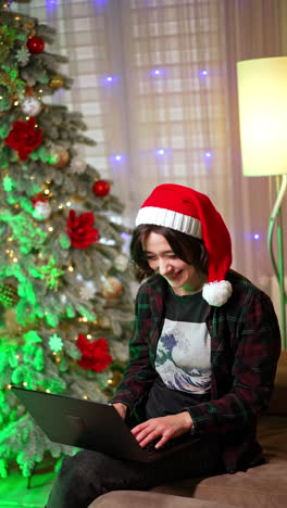 laughing caucasian girl in santa cap sitting on the sofa looks at laptop. happy woman works at home at christmas season. vertical video.