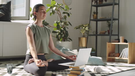 woman meditating in lotus pose with online yoga class on laptop