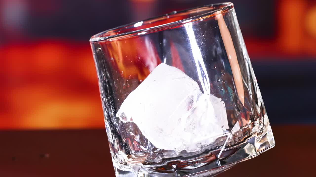 A close-up of an ice cube settling inside a tilted glass against a warm, blurred background.