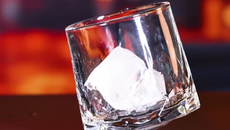 a close-up of an ice cube settling inside a tilted glass against a warm, blurred background.