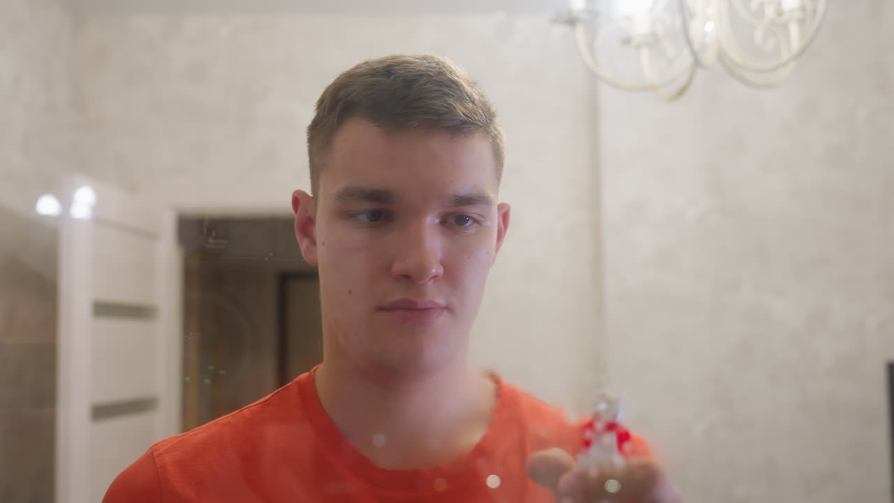 Young man in orange shirt spraying cleaning solution on bathroom mirror while focusing on removing streaks and water spots, showing household care