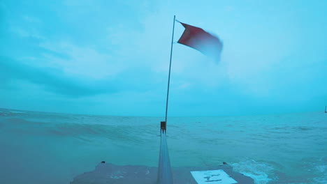 at the edge of the great lakes, a red flag waves in the wind. centered in the frame, the flagpole stands and the water crashes into their pier.