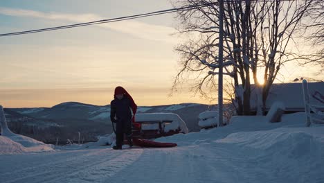 joven con sombrero rojo largo bajando del trineo y corriendo hacia atrás con el cielo naranja tranquilo de la puesta de sol en el fondo en noruega