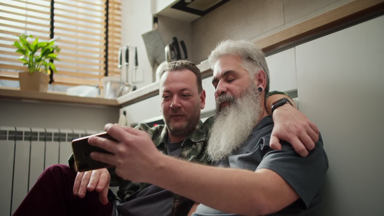 A happy elderly man with gray hair and a lush beard with his brunette boyfriend with stubble in a green checkered shirt communicates via video conference and his relatives in a modern apartment in the kitchen while sitting on the floor