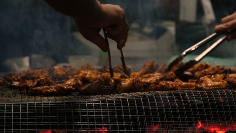 Vista-De-Cerca-De-Asar-Alitas-De-Pollo-En-La-Parrilla-De-Barbacoa-Con-Las-Manos-Sosteniendo-Tenazas