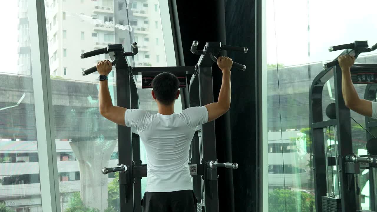 A man performs exercises on an assisted pull-up machine in a modern gym with large windows and city views