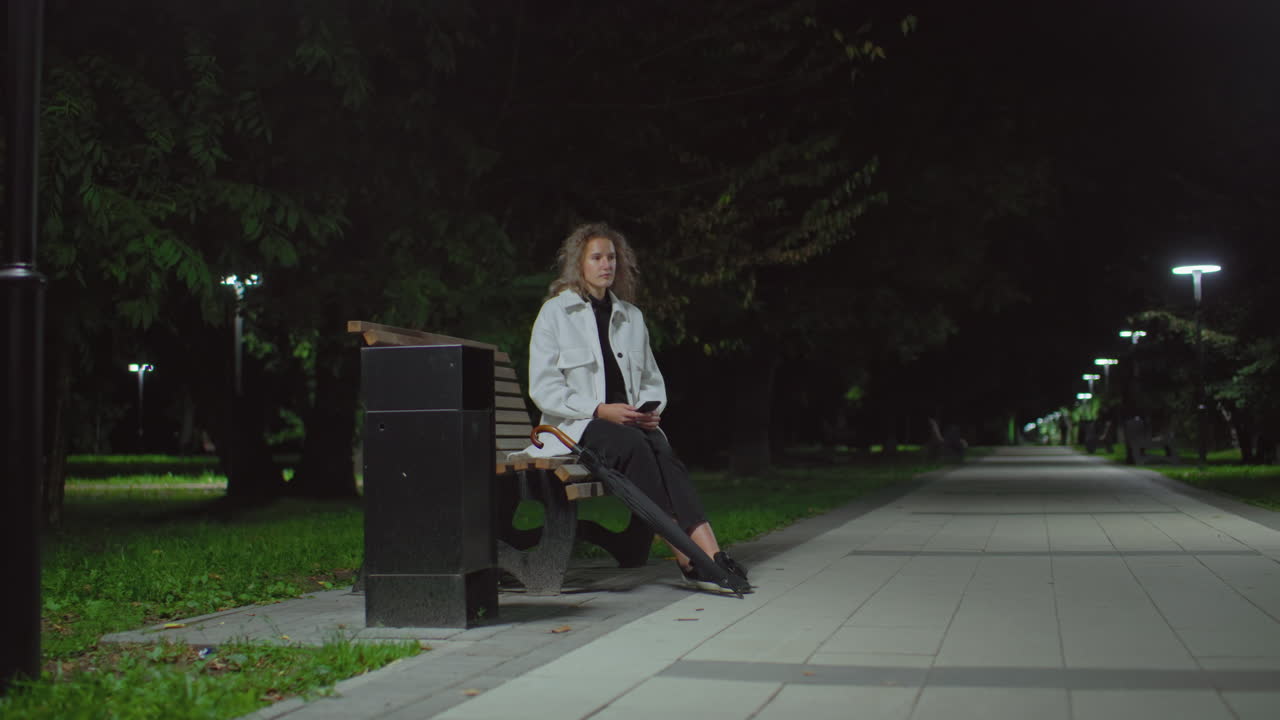 Young girl seated on bench at night using phone lifts head thoughtfully while surrounded by dim park lights, trees, and empty pathway, dressed in white coat with umbrella resting beside her