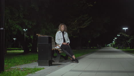 young girl seated on bench at night using phone lifts head thoughtfully while surrounded by dim park lights, trees, and empty pathway, dressed in white coat with umbrella resting beside her