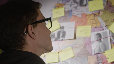 caucasian man looking at wall map with strings, stickers and photos, searching connections