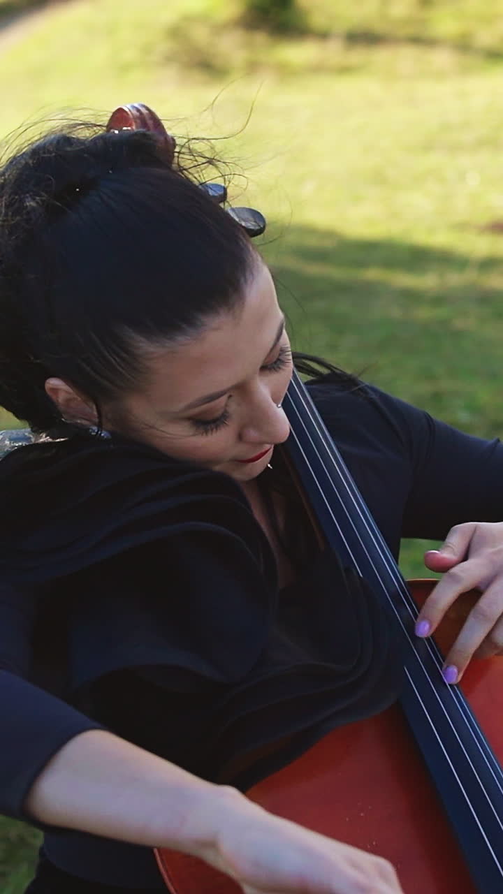 Lady wearing black dress, long earrings and red lipstick playing cello outdoors. Female musician performing music and enjoying the process. Nature backdrop. Vertical video