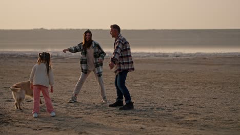 A-brunette-girl-wearing-a-hat-in-a-green-checkered-shirt-plays-with-her-large-cream-colored-dog-along-with-her-husband-and-little-daughter-while-vacationing-outside-the-city-on-a-deserted-seashore-in-summer