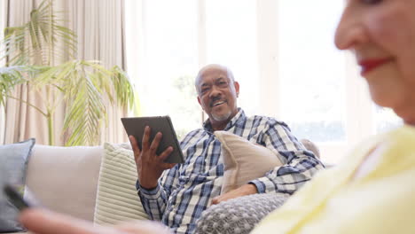 Glückliches,-Vielfältiges-Seniorenpaar-Mit-Smartphone-Und-Tablet,-Lächelnd-Im-Sonnigen-Wohnzimmer,-Zeitlupe