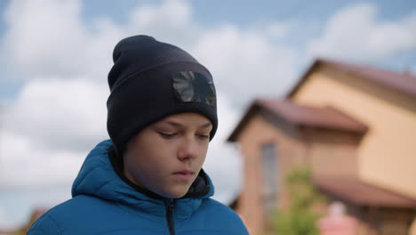 primer plano de un niño con chaqueta azul y gorra negra paseando al aire libre pensativo, fondo borroso de vegetación, casa de ladrillo y cielo suave de otoño