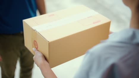 close up parcel in hand of blue uniform delivery man to customer front of the house with good service from shopping online. courier man delivering a cardboard box postal package to destination.