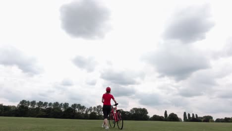 Caucasian-female-cyclist-in-red-top-walking-with-her-bike-in-a-park