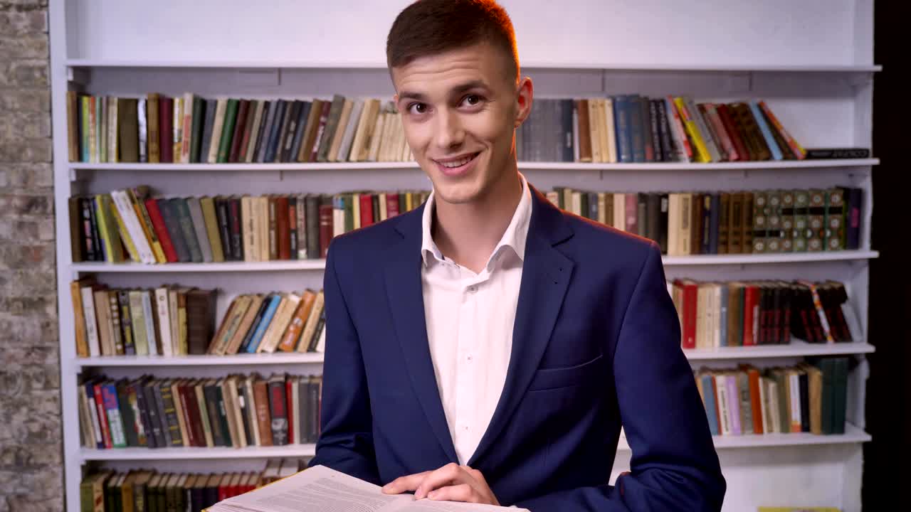 joven empresario atractivo está leyendo un libro, mirando a la cámara, sonriendo, estantería en el fondo