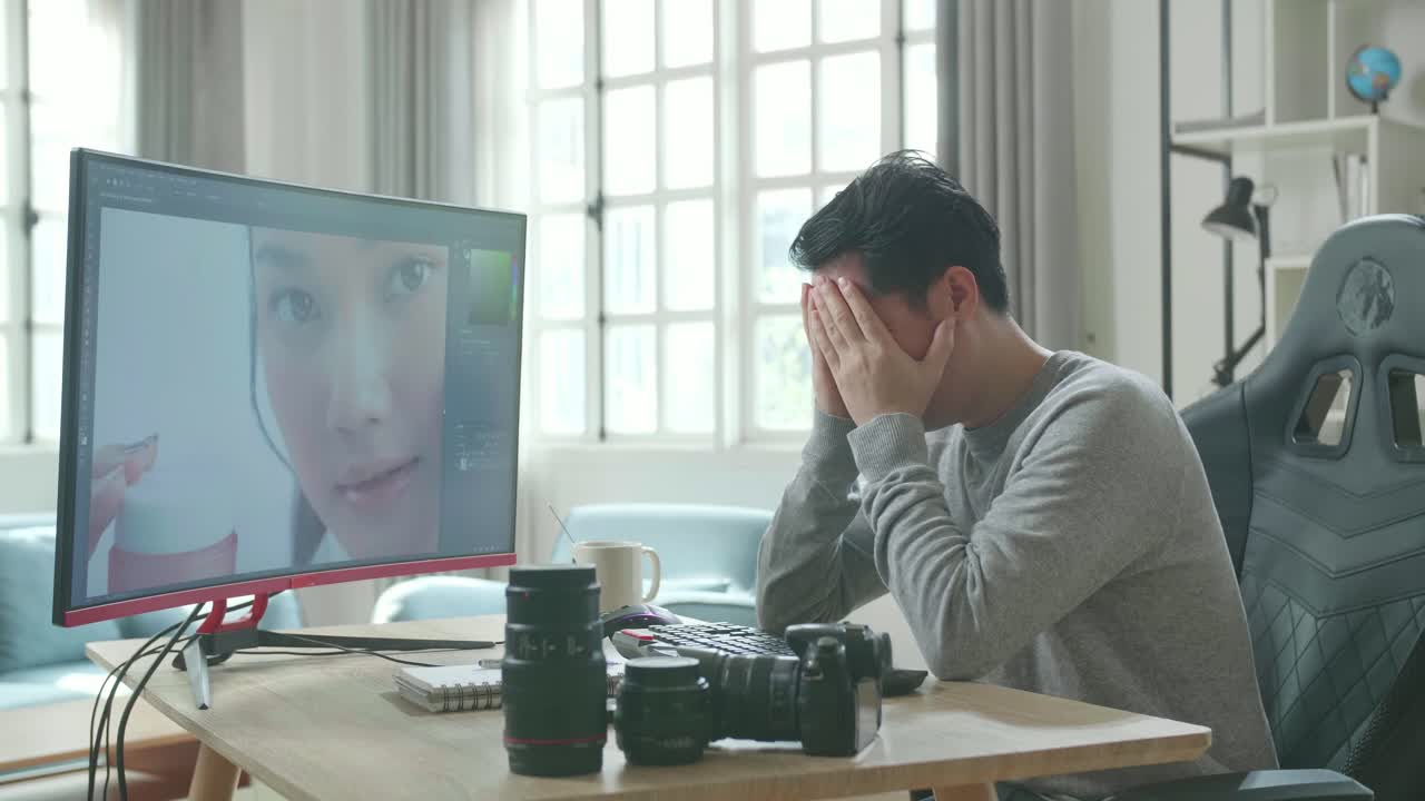 Asian Cameraman In Long Sleeved T-Shirt Being Tired While Using Desktop Computer For Editing Photo At Home.