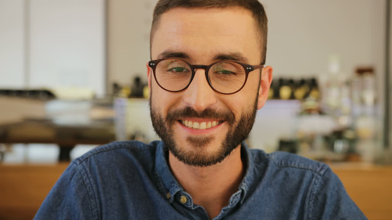 retrato de un joven con barba y gafas mirando a la cámara y sonriendo en un café
