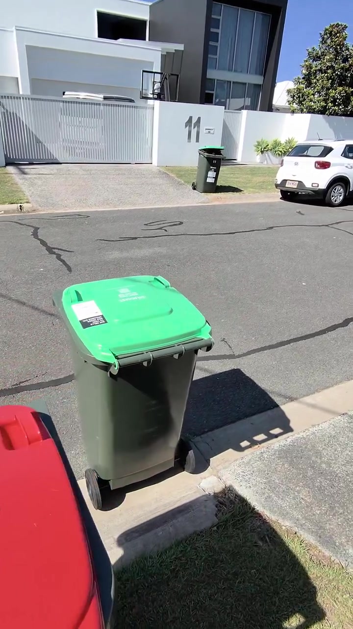 Point-of-view of man dragging red and green wheelie bins from sunny suburban curbside