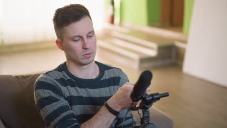 man adjusting video camera with microphone attached in creative workspace. focused on preparing equipment, surrounded by unpacked bags and modern furniture, ready for filming or content creation