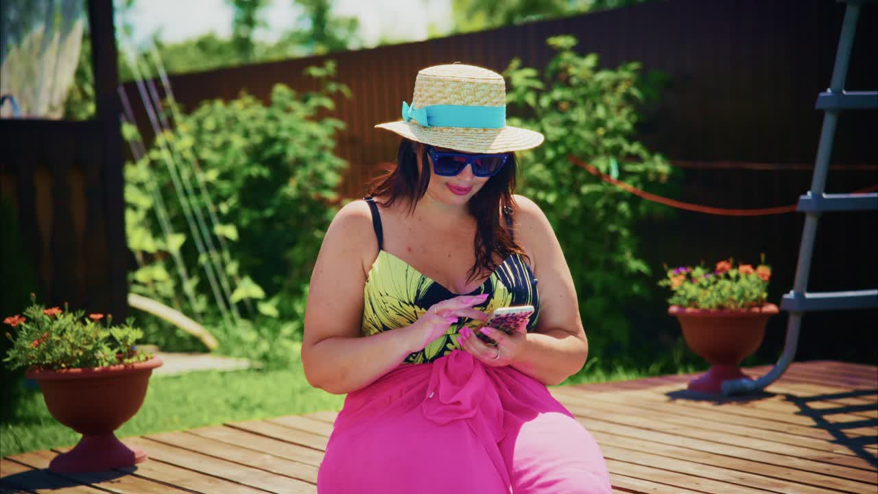 Woman in colorful swimsuit and hat relaxing outdoors while using smartphone in summer garden