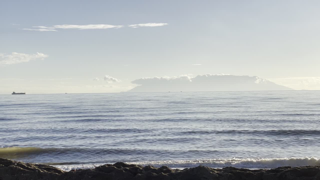 Tranquil seascape featuring gentle ocean waves under a bright sky, with a cargo ship and an island visible on the horizon, perfect for relaxation or maritime themes