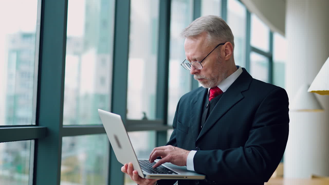 Successful businessman in glasses with a laptop in the office. Elegant mature man in suit standing near the window and holds a modern laptop.