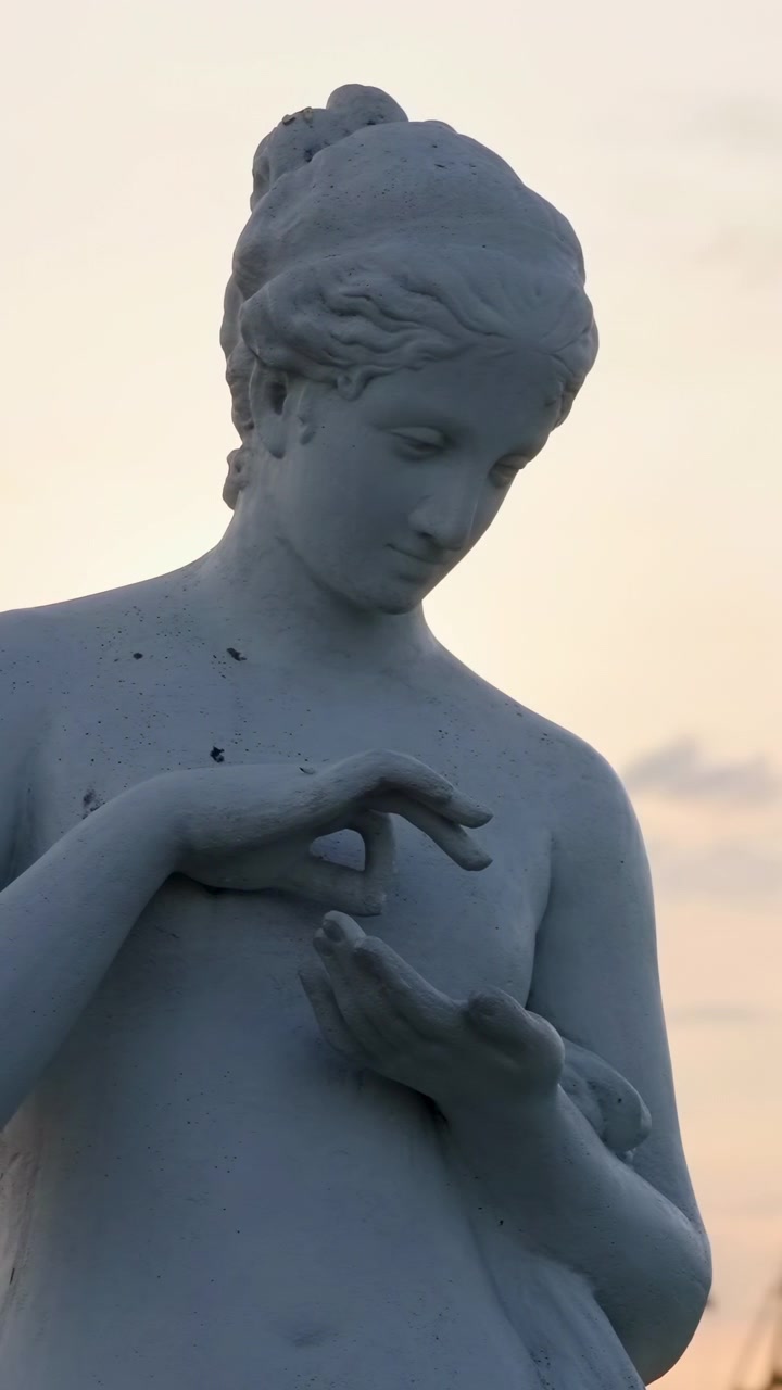 Statua di marmo al tramonto, primo piano di figura femminile, mani che formano un gesto delicato, cielo dai colori pastello tenui, in silhouette.