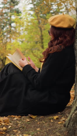 vista lateral de una mujer joven hojeando un libro en el bosque de otoño, deteniéndose con la mano en la página, sentada contra un árbol, disfruta de la lectura y la soledad mientras las hojas doradas cubren el suelo del bosque