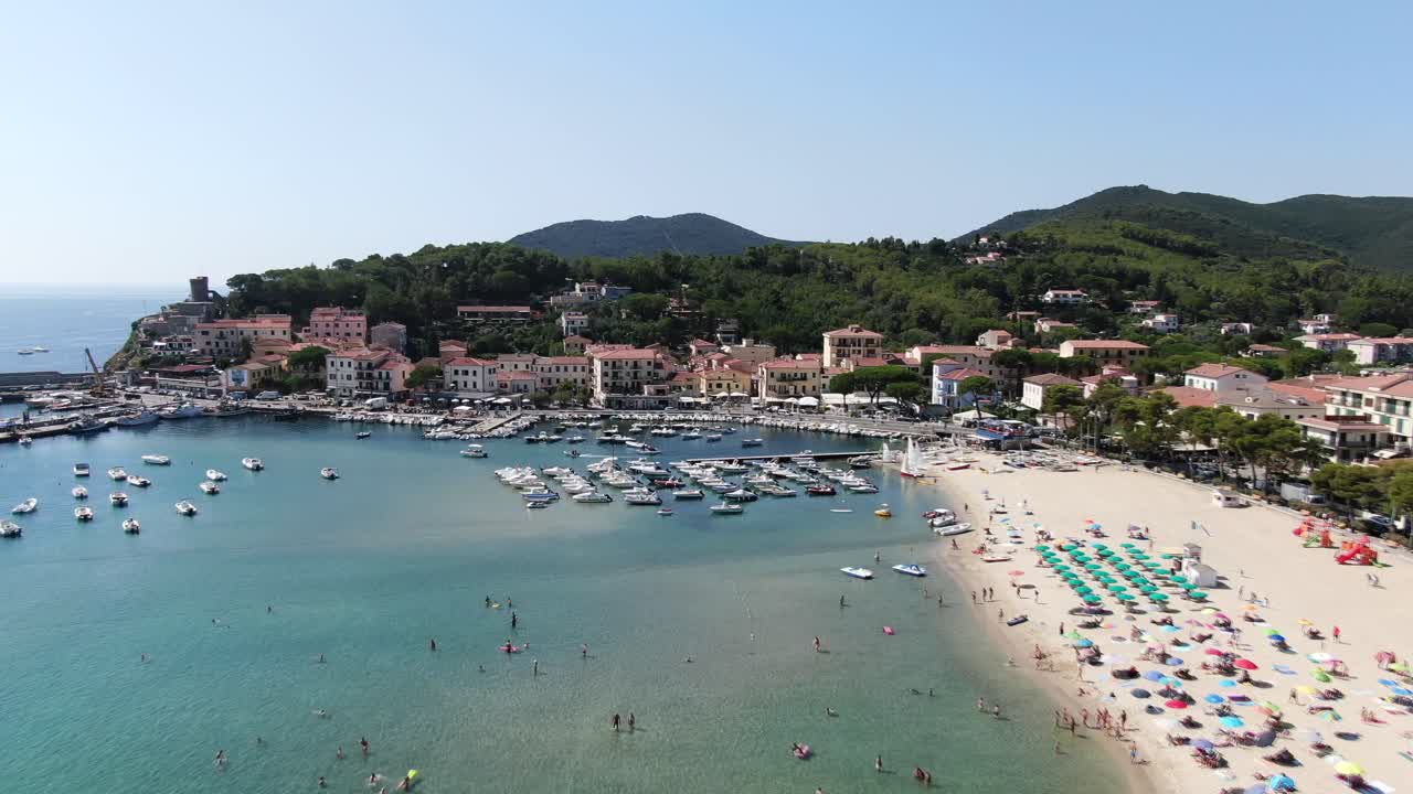 Splendida ripresa aerea di una spiaggia affollata dell'Elba con sabbia incontaminata, onde scintillanti e barche ancorate al largo