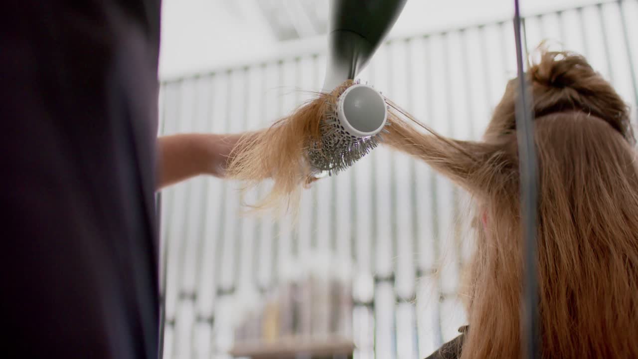 Caucasian female hairdresser styling client's long hair with hairdryer and brush, in slow motion