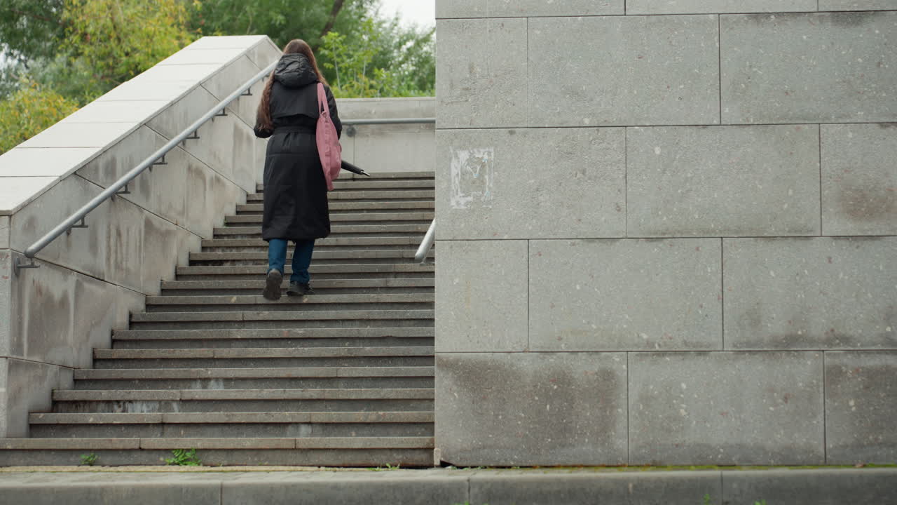 woman walking up stone stairs in urban park overcast sky wearing dark coat and backpack with pink bag ascending concrete staircase past lamppost and greenery quiet routine commute capturing solitary