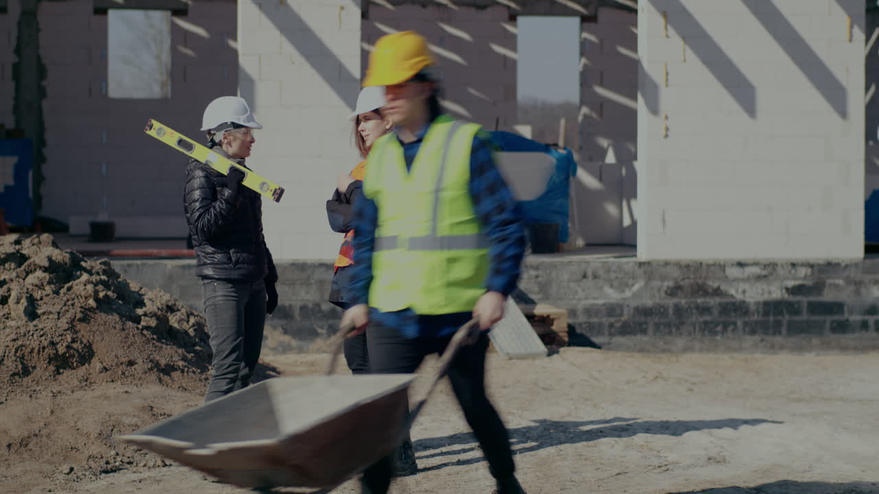 Young female engineer discussing with worker while male coworker pushing wheelbarrow at construction site