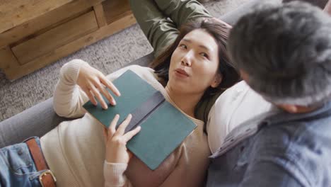 casal feliz e diversificado sentado na sala de estar com um livro e conversando juntos