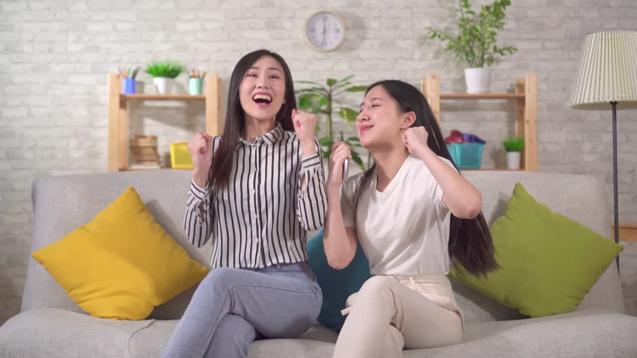 두 명의 행복한 아시아 소녀들이 거실의 소파에 앉아 스마트폰을 보고 승리한 것에 행복합니다.