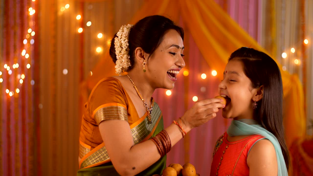Smiling Indian mother feeding sweets to her young daughter during festivities - Happy Family