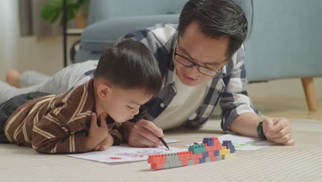 close up of asian father and son lying on the floor in the room with plastic toy brick drawing together at home