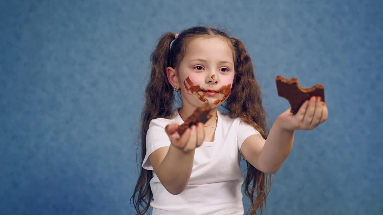 Dirty face of a girl eating chocolate. Two pieces of sweet chocolate in child's hands. Portrait of a little kid with smeared face isolated in studio.