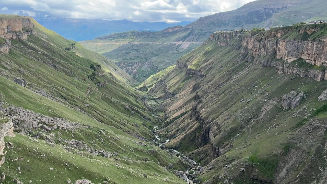 Una vista aerea si sposta lentamente verso l'alto rivelando un fiume serpeggiante sul fondo di una profonda valle di montagna verde con pendii ripidi e rocciosi sotto un cielo nuvoloso in Dagestan.
