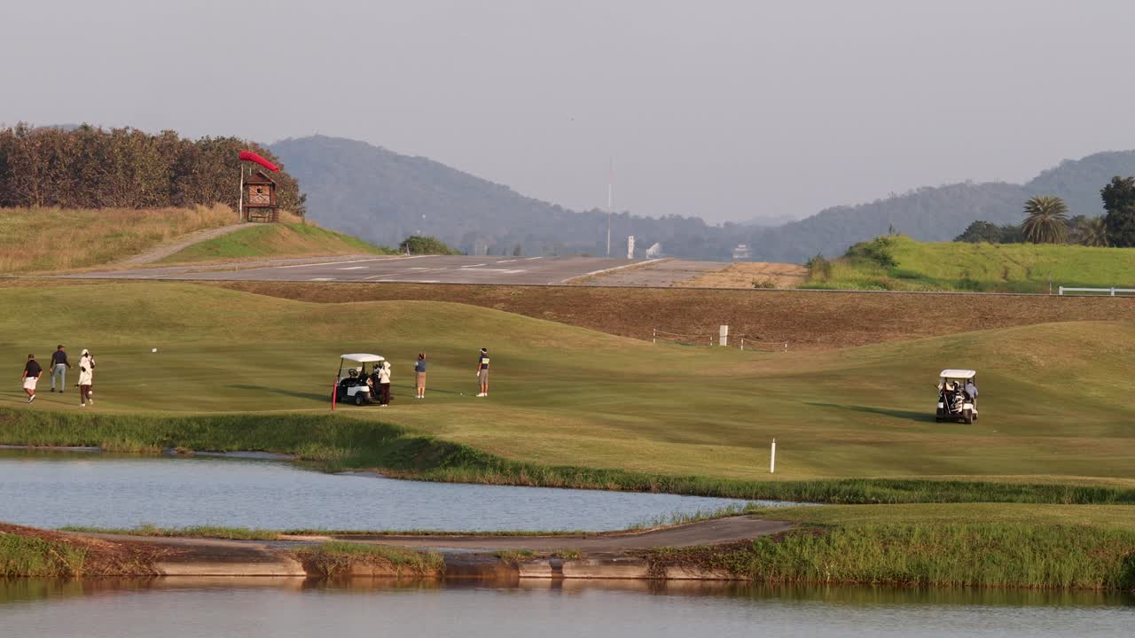 Golfers and carts traverse lush fairway near lake, mountains, and sunset, wide static shot