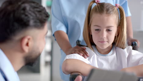 Biracial-doctor-talking-to-caucasian-girl-patient-sitting-in-wheelchair-in-hospital,-slow-motion